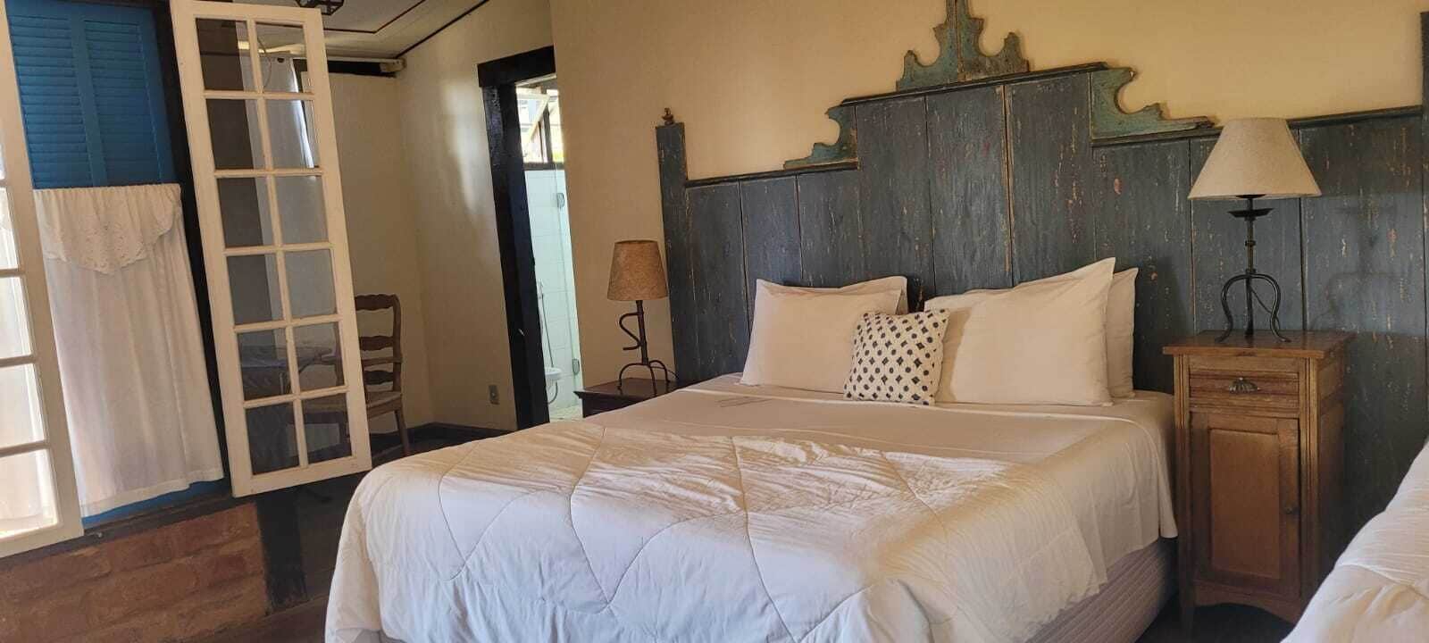 Un dormitorio con una cama grande, un cabecero de madera, una mesita de noche con lámpara y una puerta que da a otra habitación.