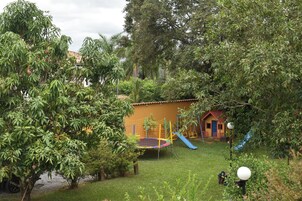 A playground with a slide and climbing net, surrounded by lush greenery and a small building.