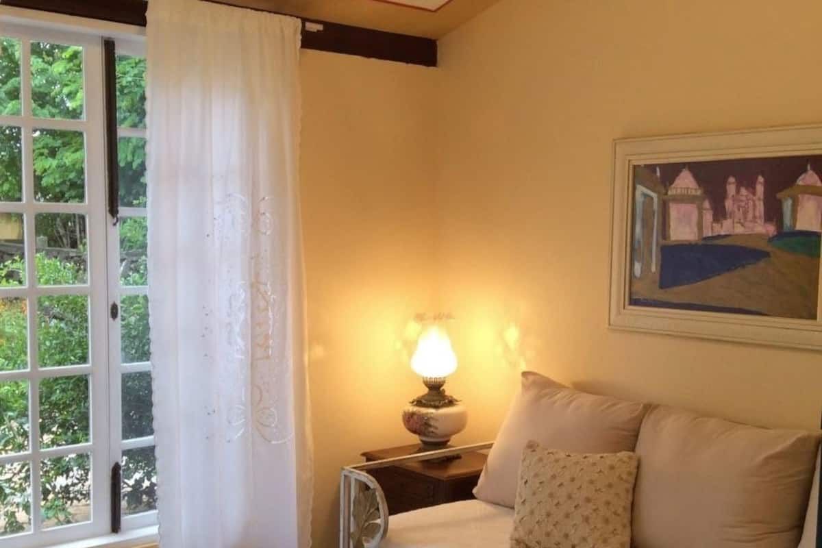 Ein gemütliches Zimmer mit einem Bett, einem Nachttisch mit Lampe, einem Fenster mit Vorhängen und einem Bild an der Wand.