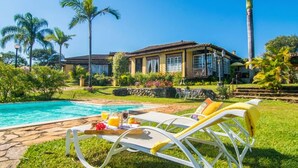 Un área junto a la piscina con sillones de descanso y una mesa con alimentos para el desayuno, rodeada de exuberante vegetación y una amplia casa con techo de tejas.