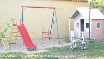 Un parque infantil con columpios y un tobogán, un edificio pequeño y una escalera.
