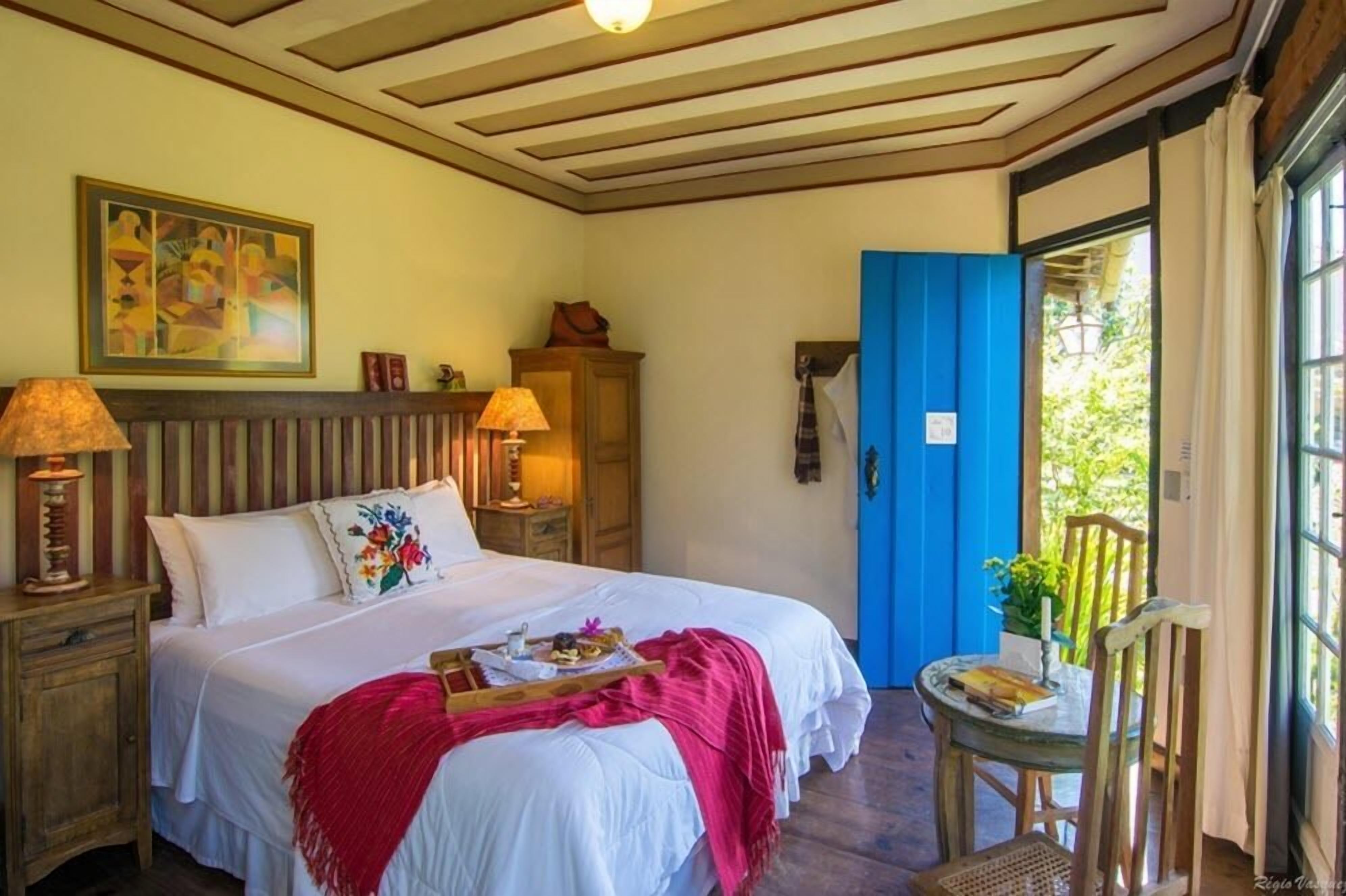 Quarto com cama grande, abajures, porta azul e vista para o verde lá fora.