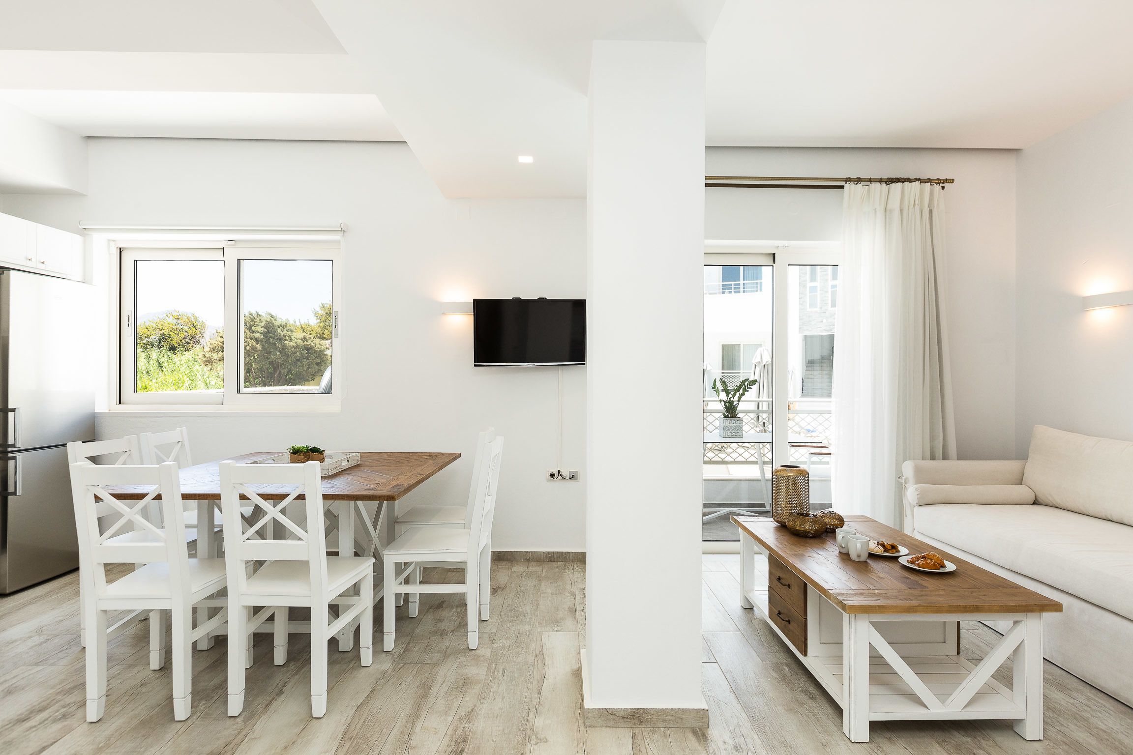 A modern living room with a wooden dining table, white chairs, a sofa, and a television mounted on the wall.