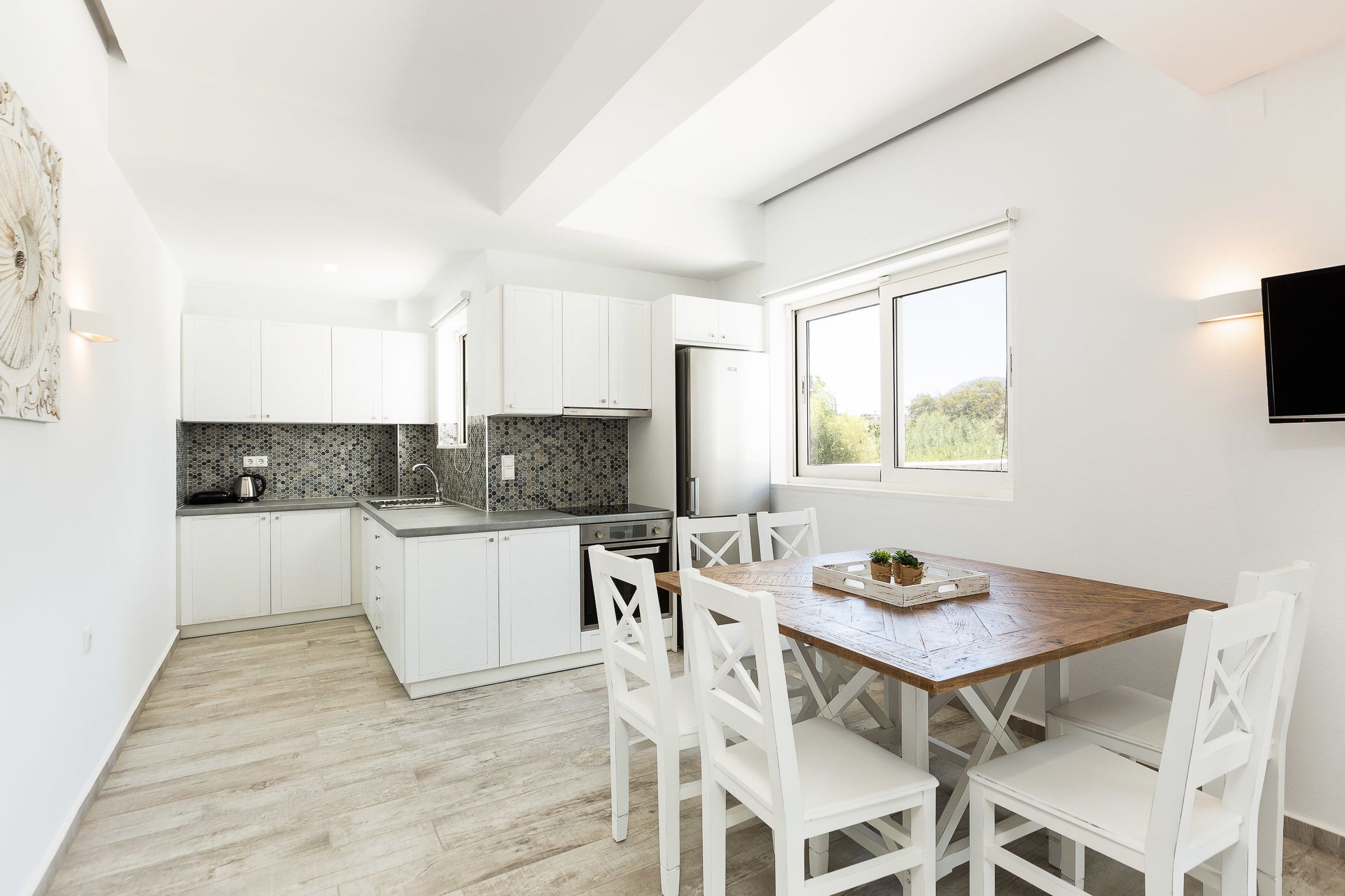A modern kitchen with white cabinets, a wooden dining table, and a large window.