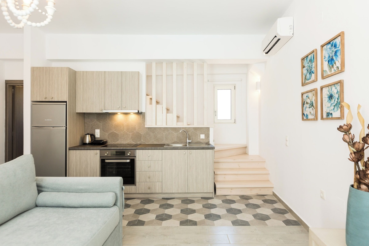 A modern kitchen with light wood cabinets, a stainless steel refrigerator, and a built-in oven. There is a staircase leading to an upper level, and a dining area with a light blue sofa.