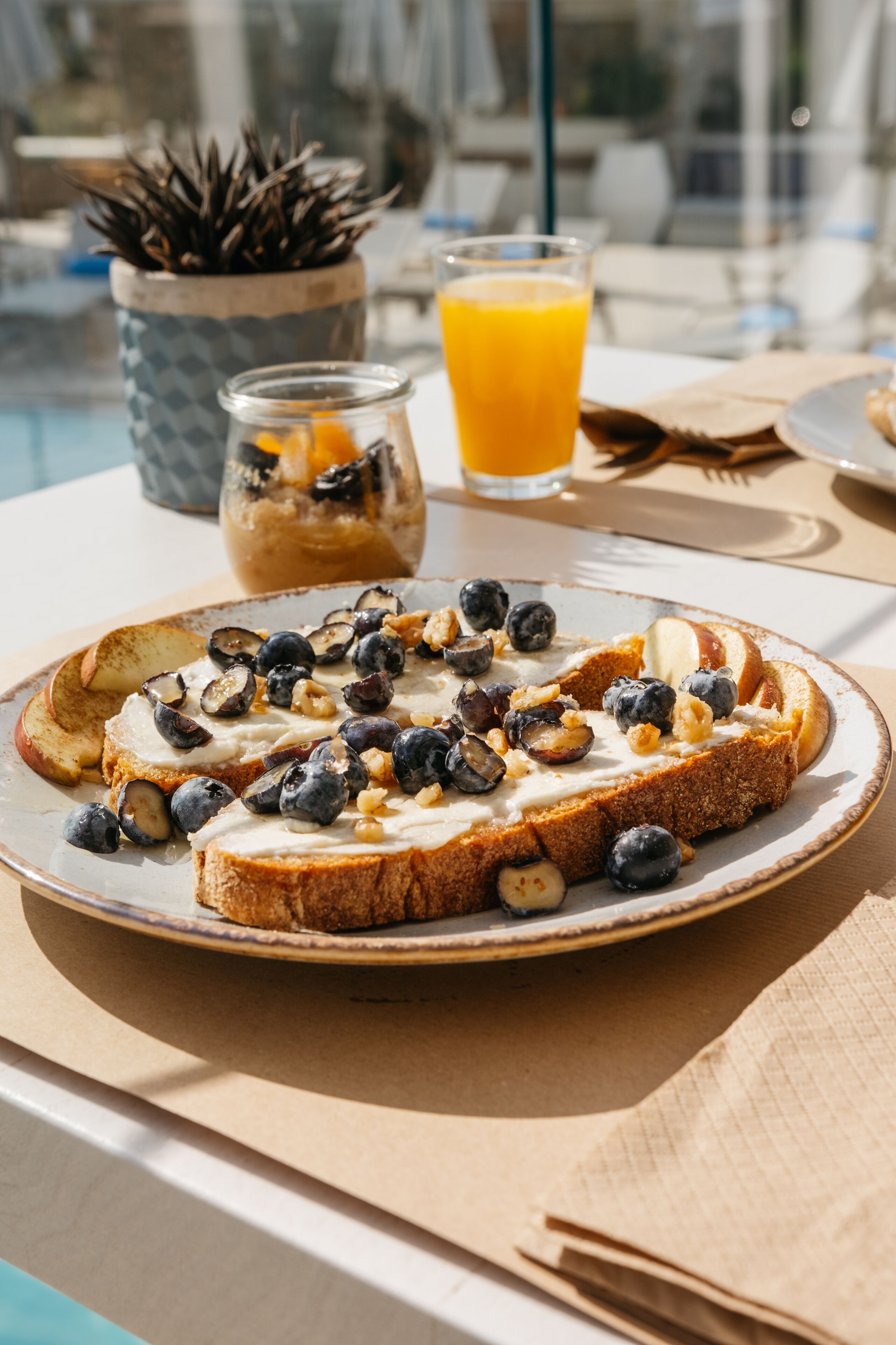 Une table mise pour le petit-déjeuner avec des tartines garnies de myrtilles et de noix, un verre de jus d’orange et un petit pot de confiture.