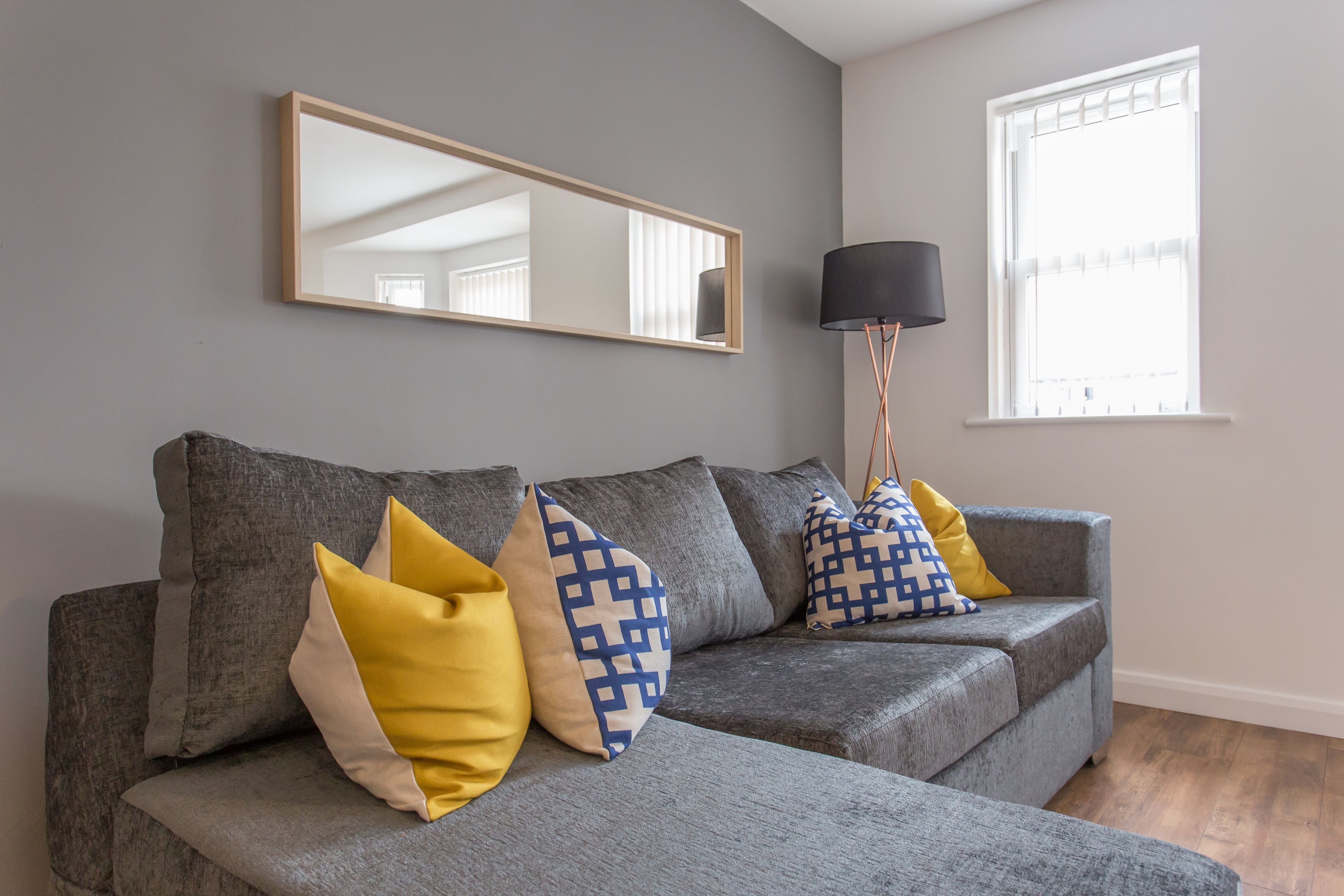 A grey sofa with yellow and blue patterned cushions, a floor lamp, and a large mirror on the wall.