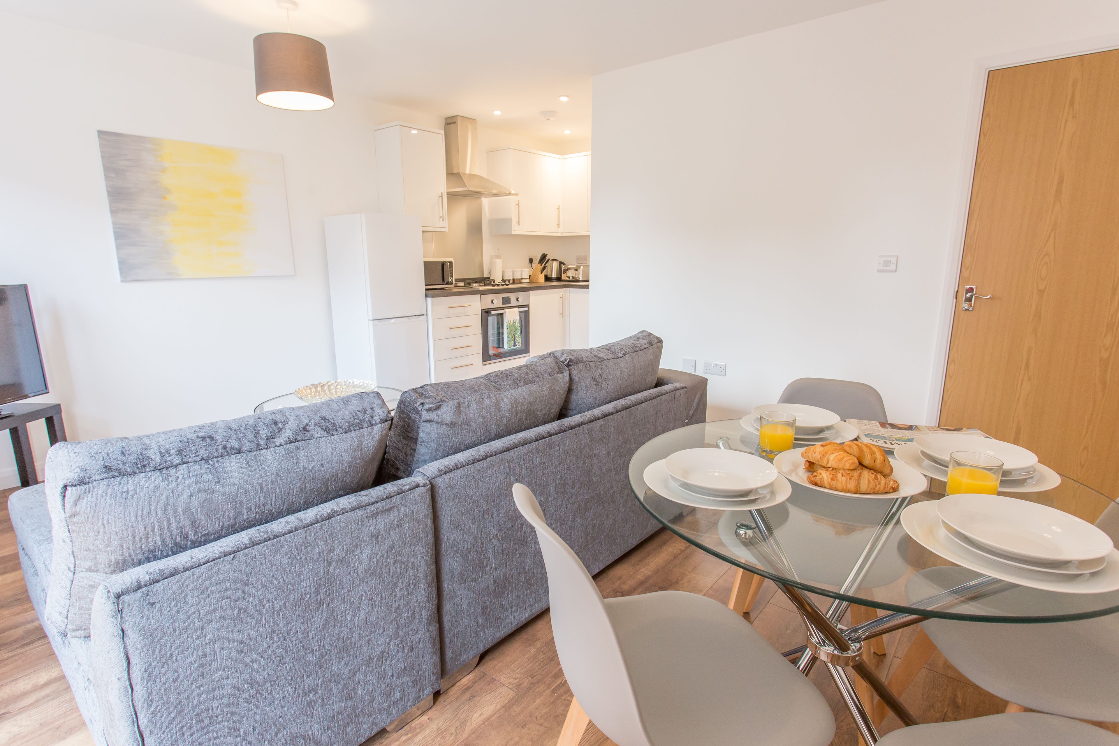 A modern living room with a grey sectional sofa, a glass dining table with plates and croissants, and a kitchen in the background.
