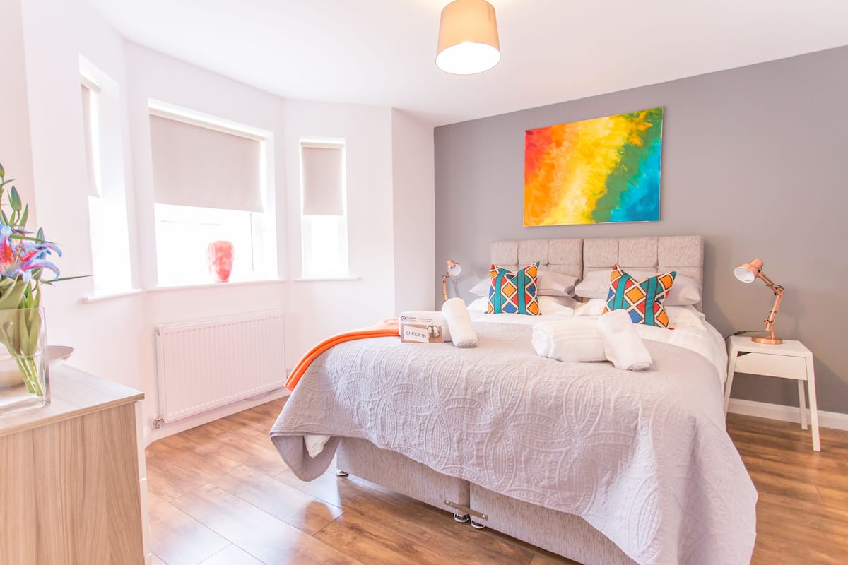 A modern bedroom with a large bed, a colorful abstract painting, and a bedside table with a lamp.