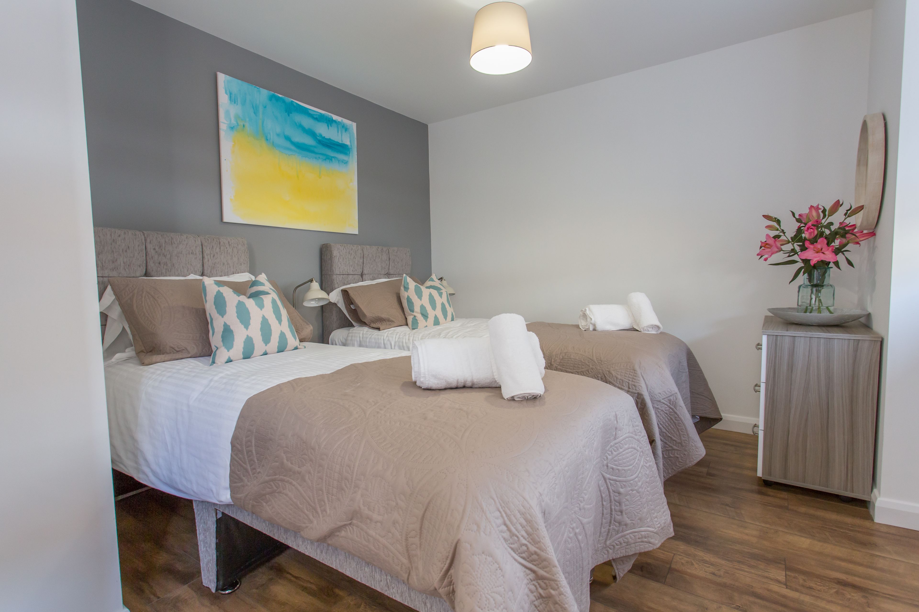 A modern bedroom with two beds, a grey headboard, a painting, a vase with flowers, and a wooden cabinet.