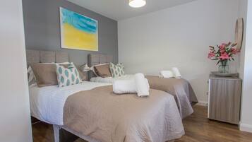A modern bedroom with two beds, a grey headboard, a painting, a vase with flowers, and a wooden cabinet.