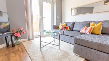 A living room with a grey sofa, a glass coffee table, a TV on a stand, and a vase of pink flowers.