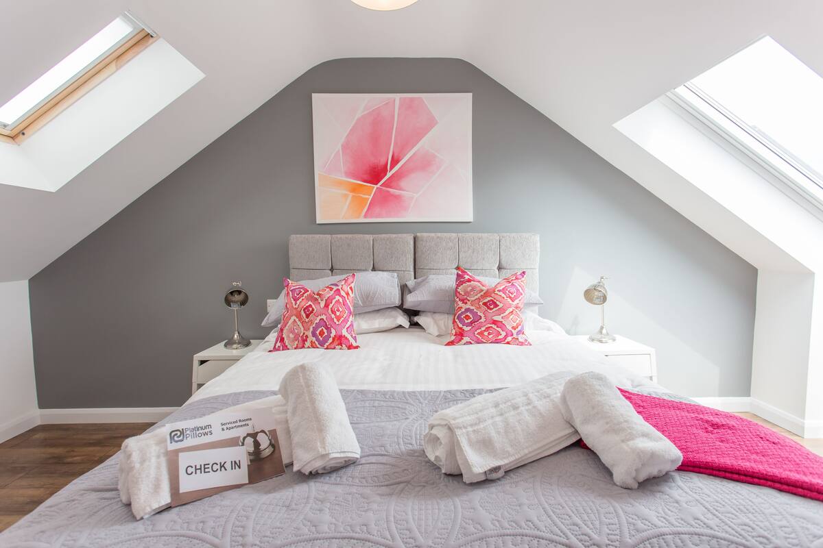 A bedroom with a sloped ceiling, a bed with patterned pillows, a bedside table with a lamp, and a check-in box on the floor.