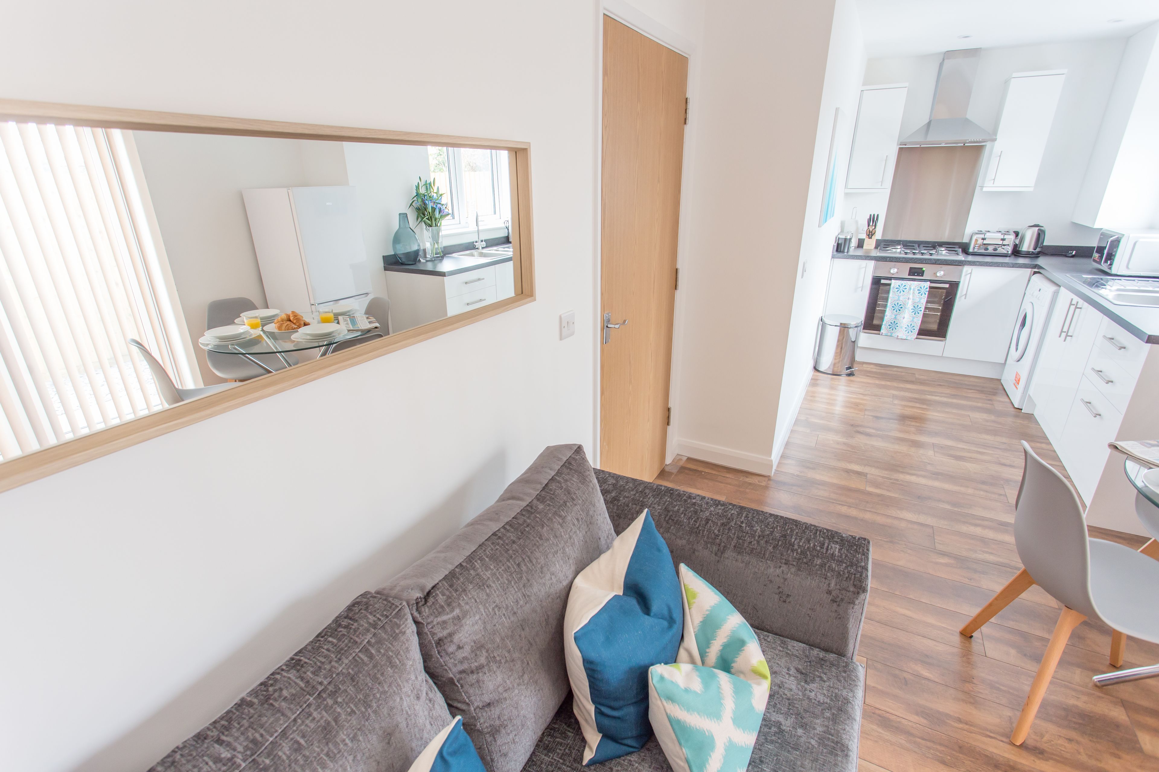Una sala de estar moderna con un sofá gris, un espejo grande y una zona de comedor con una mesa lista para el desayuno.