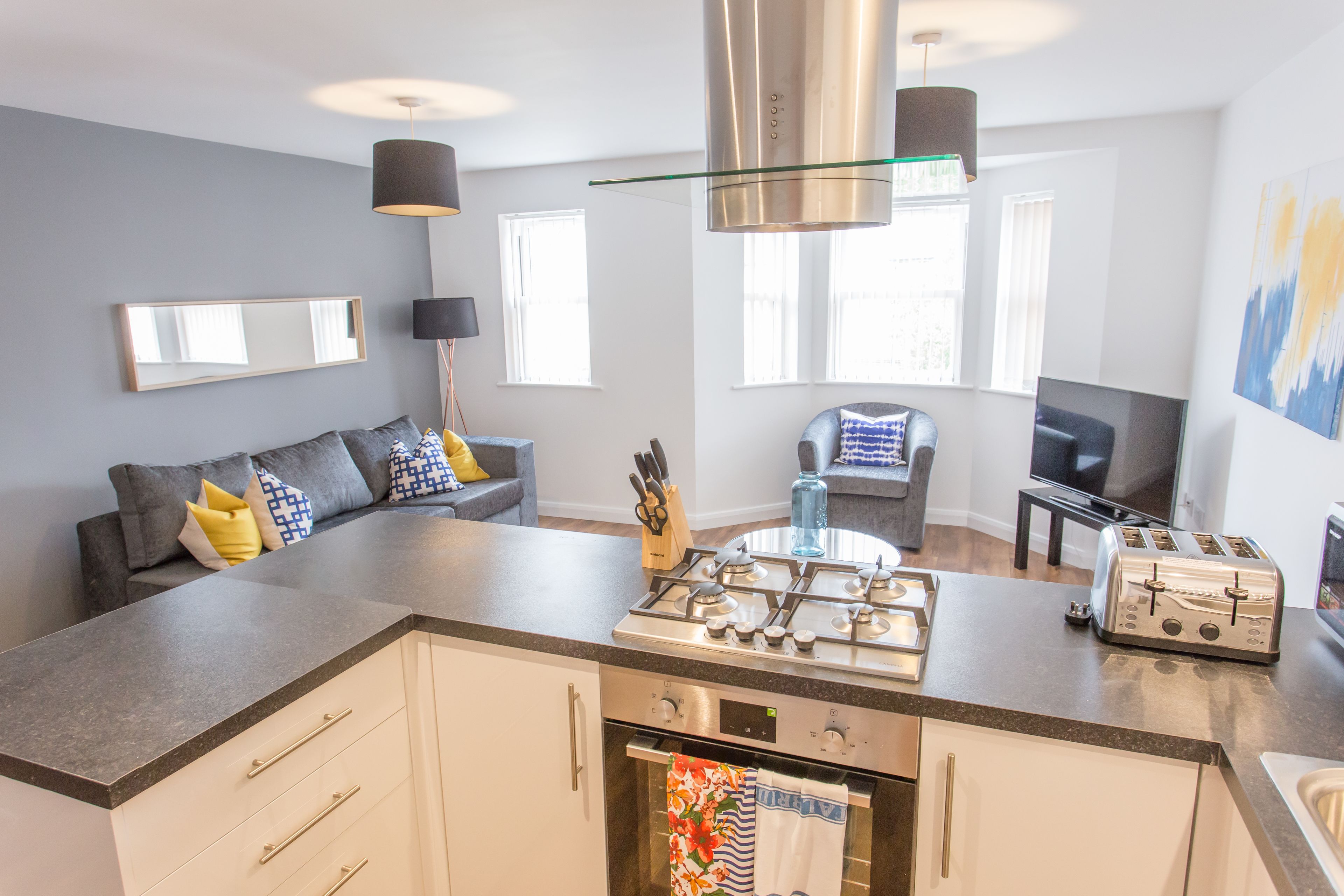 A modern kitchen with a central island, built-in oven, and a gas stove. There is a dining area with a table and chairs, and a living room with a sofa and a TV.