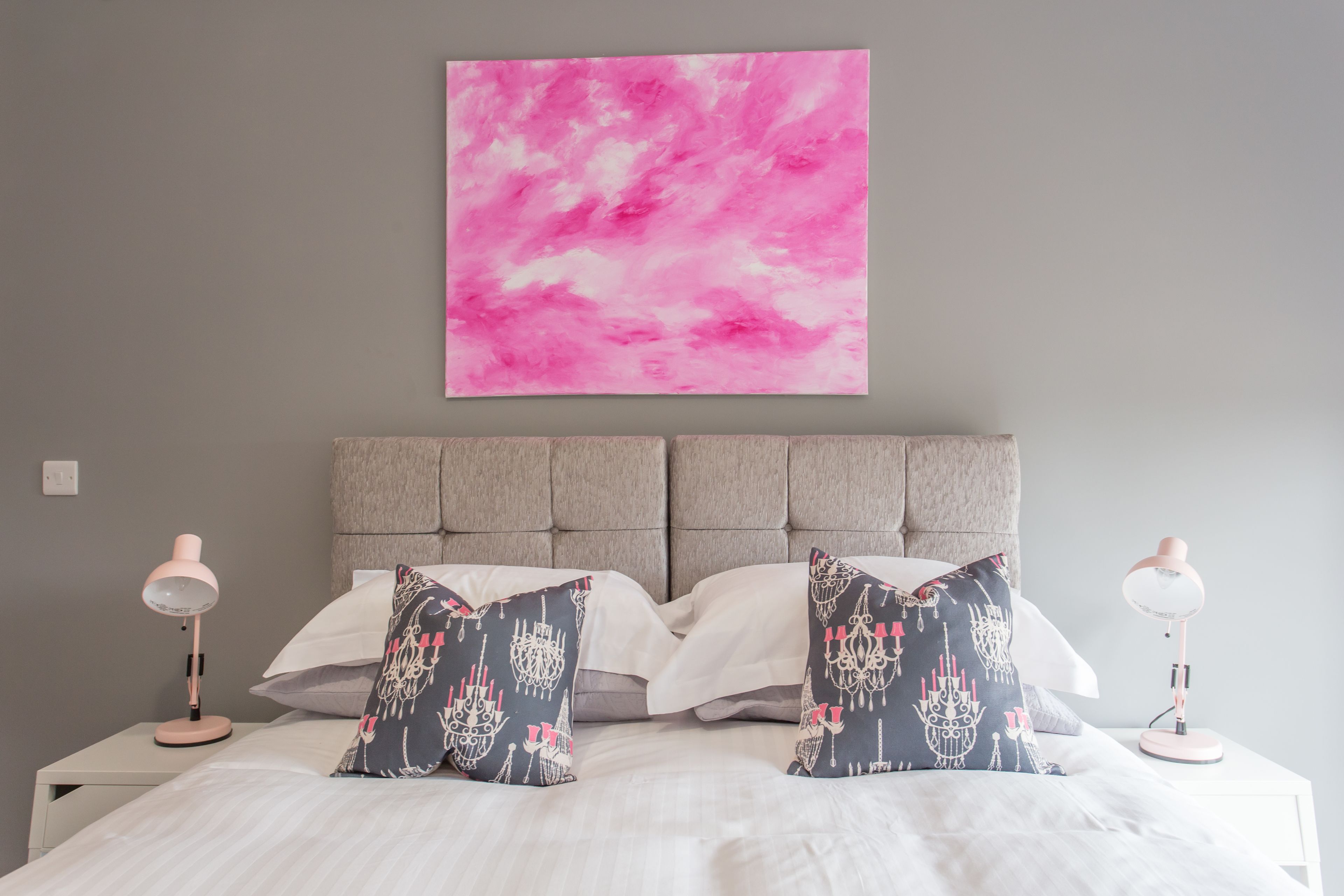 Una cama bien hecha con sábanas blancas, dos almohadones con estampado de candelabro y un cuadro abstracto rosa sobre la cama.