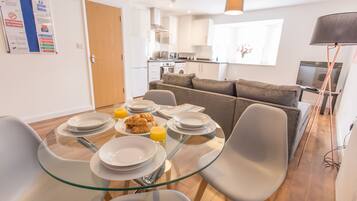 Een moderne keuken met witte kasten, een eethoek met een glazen tafel, en een grijze bank.
