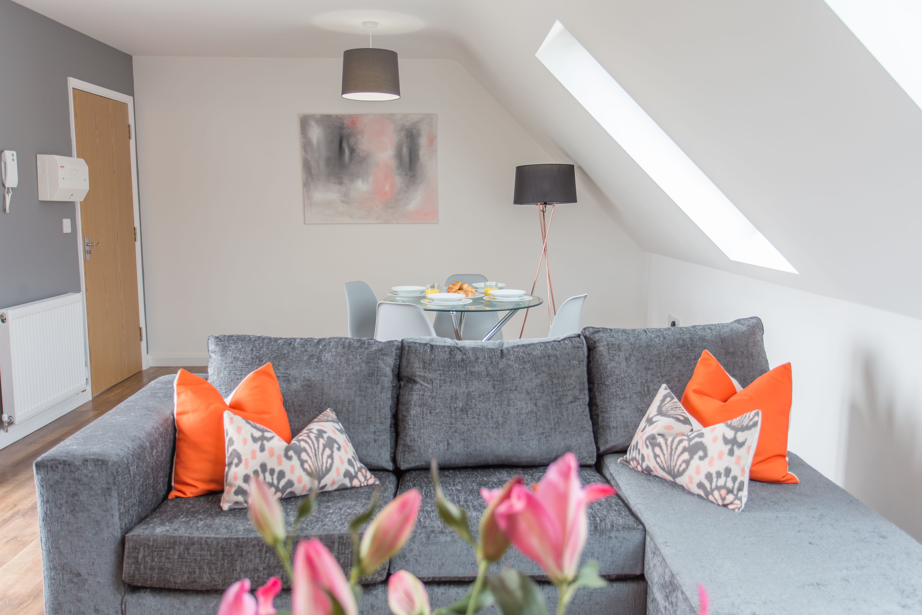 A modern living room with a grey sofa, orange and patterned cushions, a dining table with plates and glasses, and a large abstract painting on the wall.