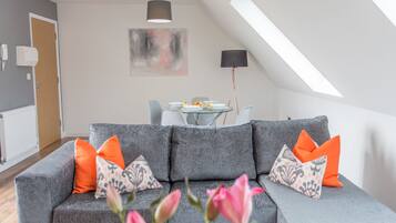 A modern living room with a grey sofa, orange and patterned cushions, a dining table with plates and glasses, and a large abstract painting on the wall.