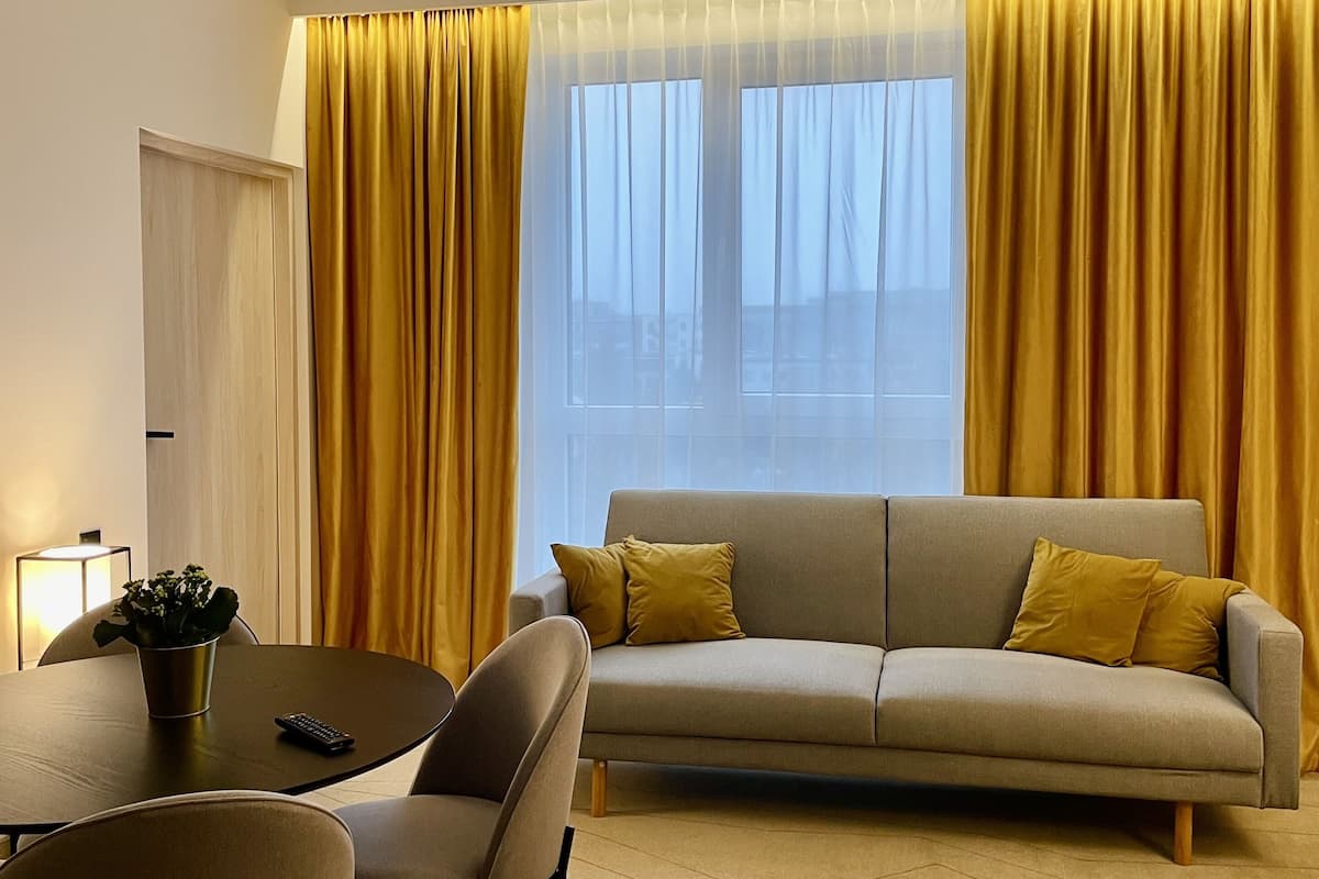 A modern living room with a sofa, two armchairs, a coffee table, and a dining table. There is a window with yellow curtains, a door, and a lamp on the table.