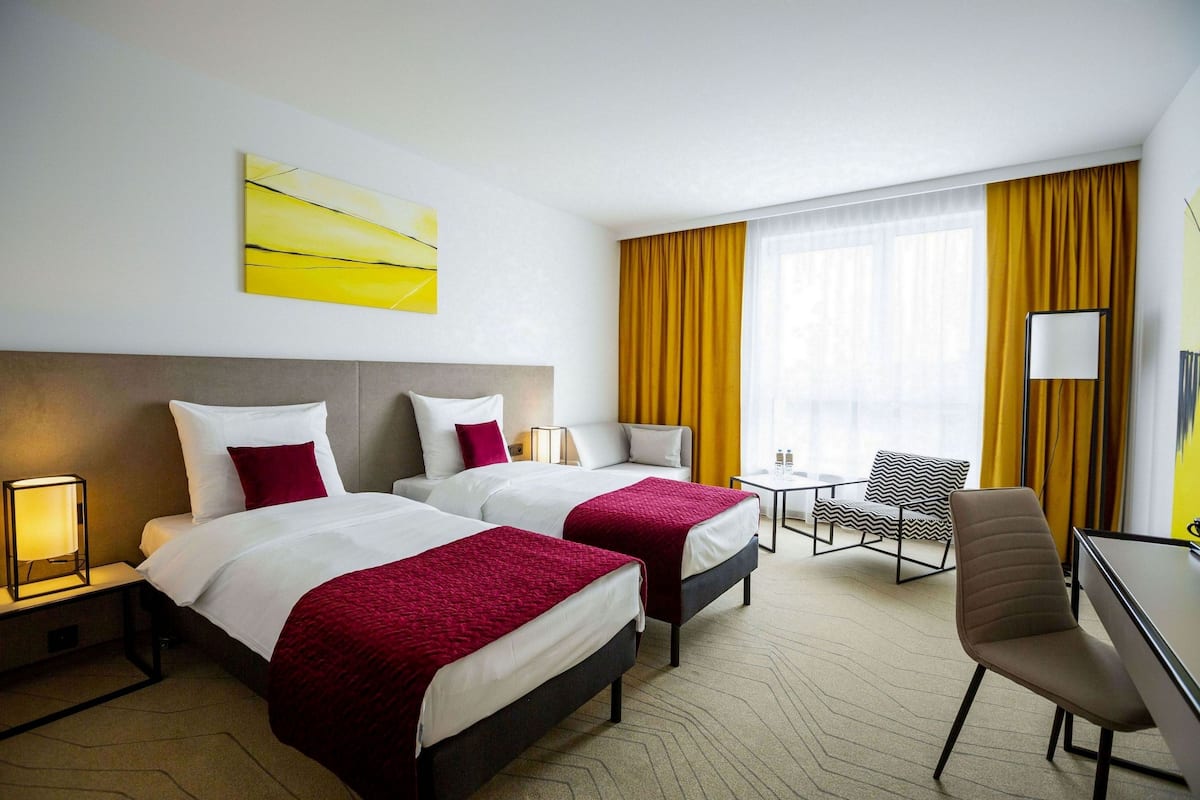 A hotel room with two beds, a desk, a chair, and a large window with curtains.