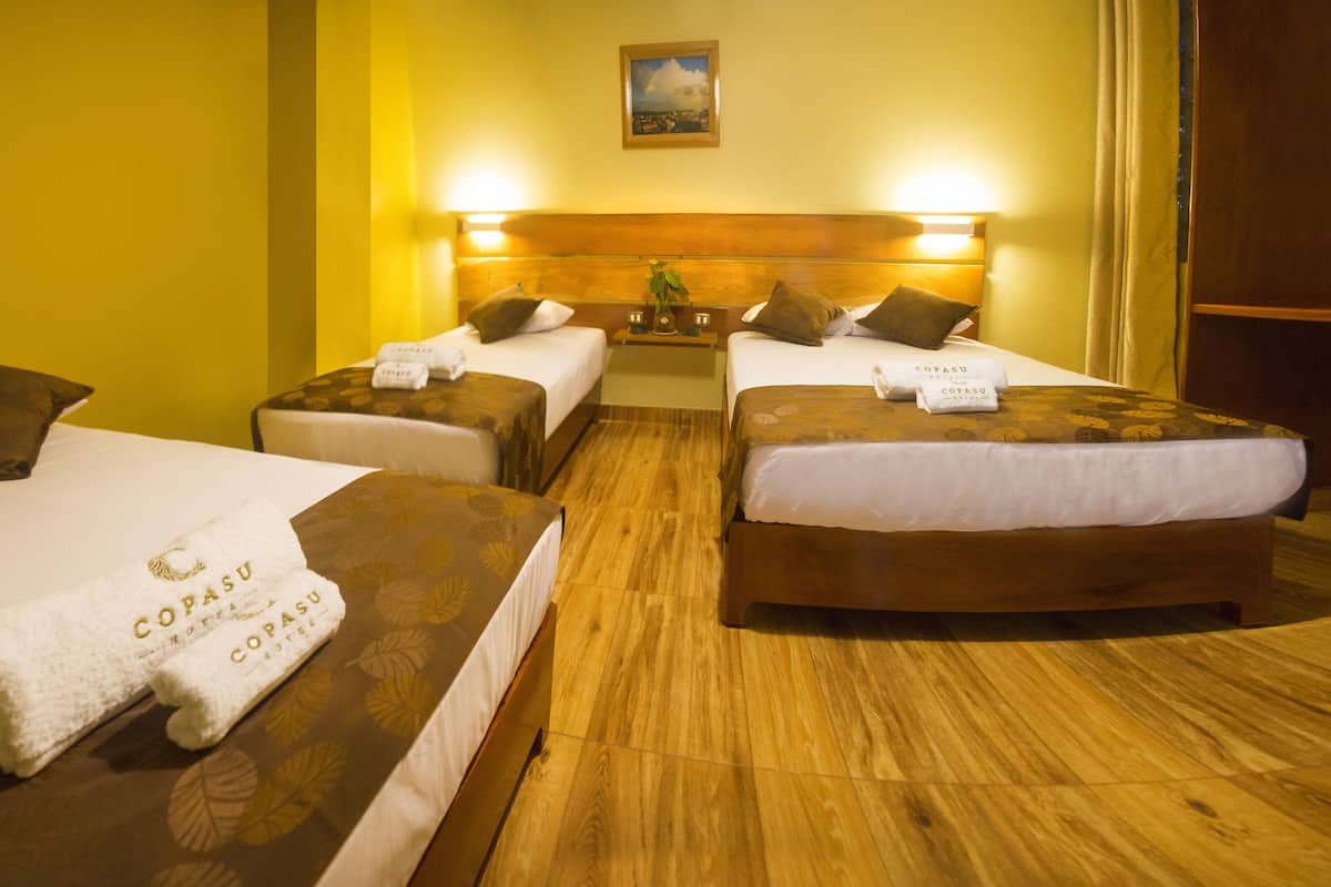A hotel room with two beds, each with a wooden headboard and a floral bedspread. There are two towels on each bed with the hotel name 'COPASU' printed on them. The room has a wooden floor and a painting on the wall above the beds.