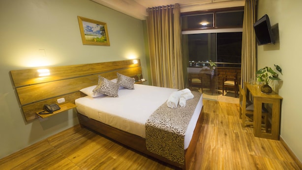Habitación de hotel con una cama de madera, una mesita de noche con teléfono, un televisor montado en la pared y un balcón con vistas a la ciudad.