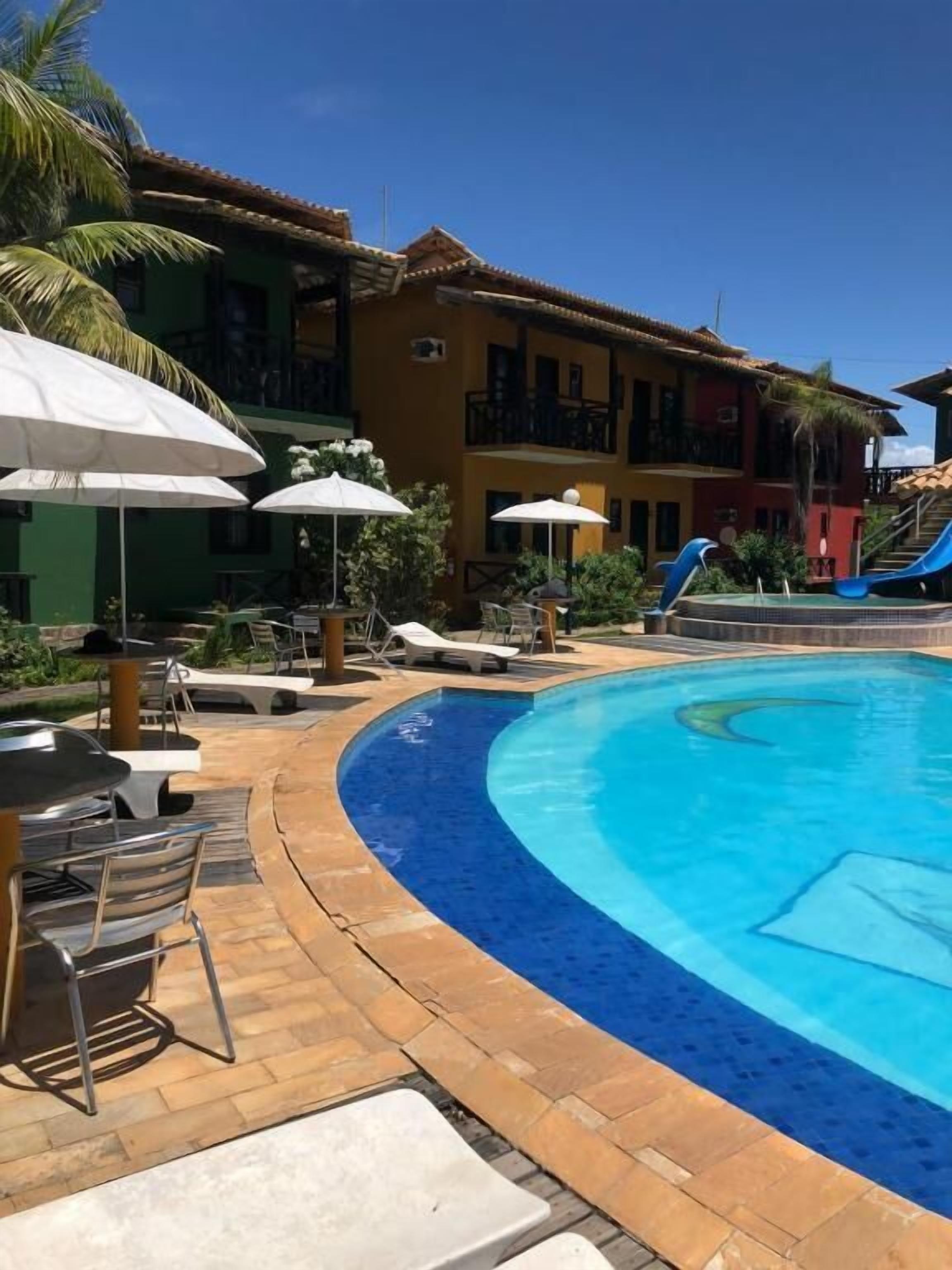 Una piscina con sillones de playa y sombrillas, un edificio con balcones y un cielo azul despejado.