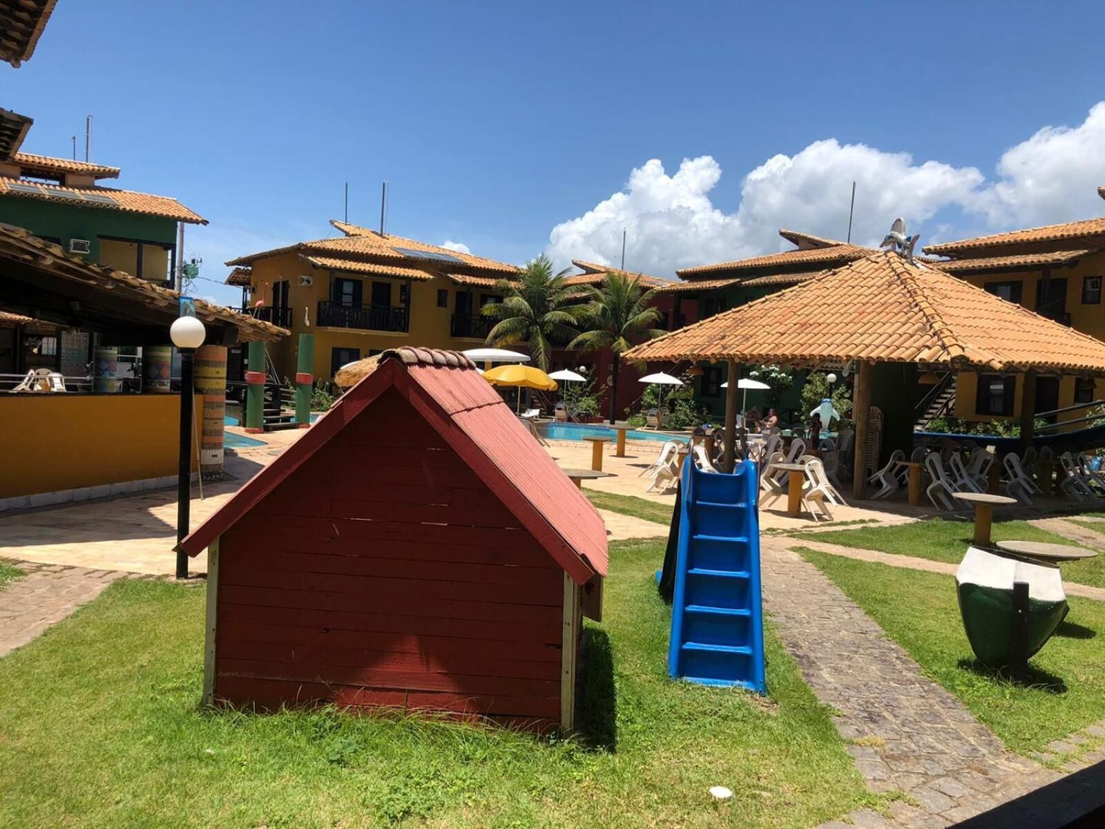 Ein Kinderspielbereich mit einem kleinen, roten Spielhäuschen, einer blauen Rutsche und einem Pool mit Sonnenschirmen.