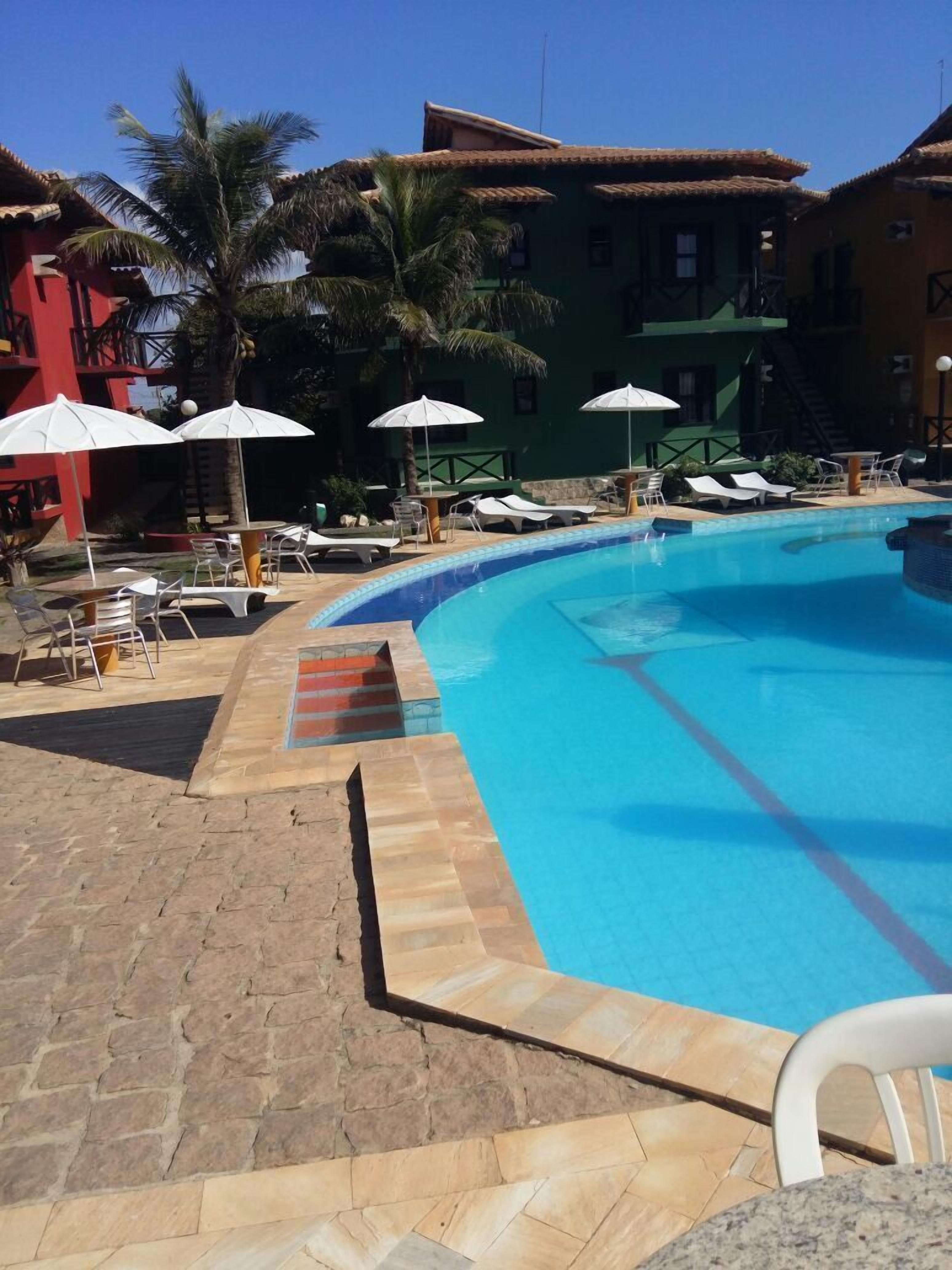 A swimming pool with lounge chairs and umbrellas, surrounded by buildings with tiled roofs and palm trees.