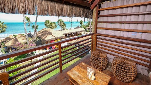A wooden balcony with a view of the beach and ocean, featuring a thatched roof, wooden railings, and wicker furniture. - Carnaubinha Praia Resort (Luis Correia)
