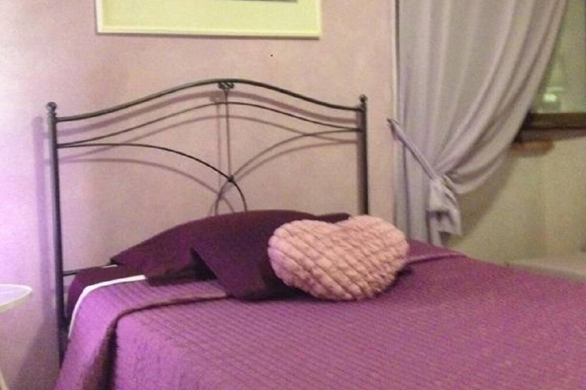 A bed with a purple bedspread and pillows, a metal headboard, a framed picture on the wall, and a window with curtains.