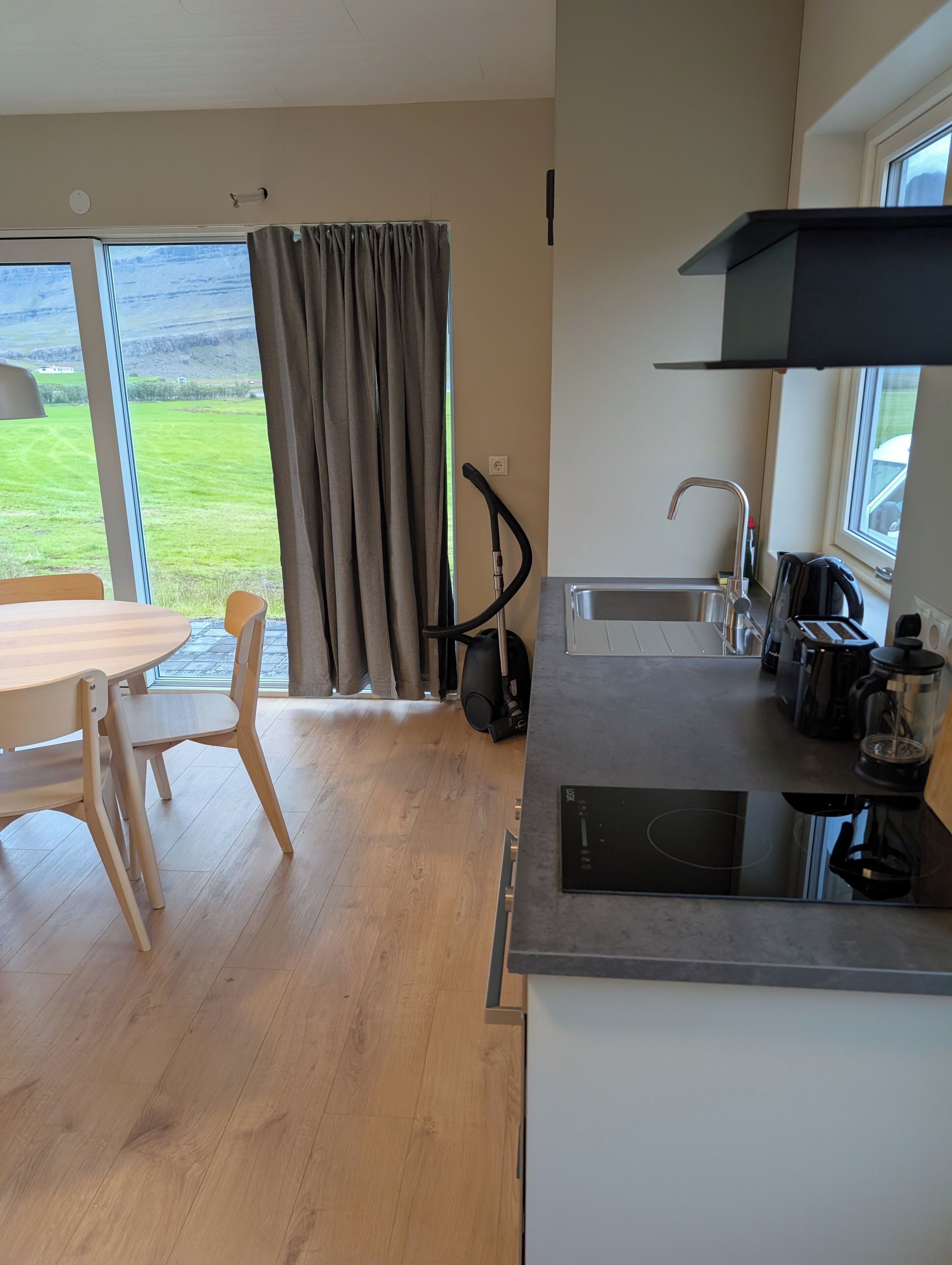 Una cocina moderna con estufa integrada, un lavamanos y una cafetera. Hay una mesa de comedor con sillas y vistas a un paisaje verde por la ventana.