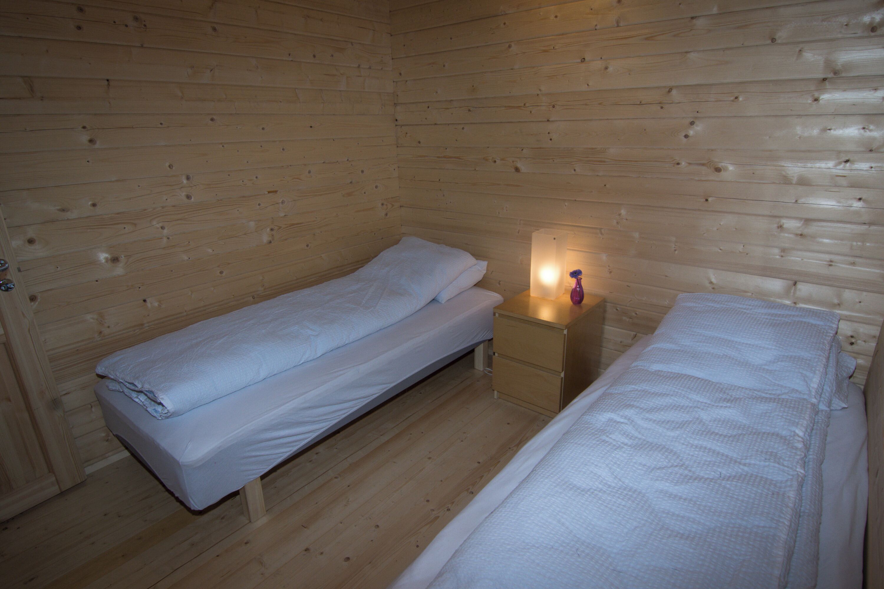 Ein kleines Zimmer in einer Holzhütte mit zwei Einzelbetten, einem Nachttisch mit Lampe und einer Holztür.