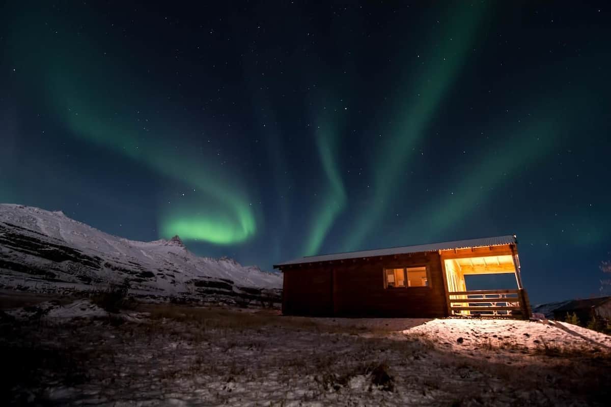 En stuga av trä med upplyst interiör, omgiven av snötäckt terräng och norrsken på natthimlen.