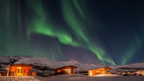 A snow-covered landscape with wooden cabins and a view of the Northern Lights. - Bragdavellir Cottages (Djupivogur)