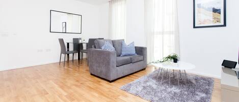 A modern living room with a grey sofa, a dining table with chairs, a small round table with a plant, and a grey rug.