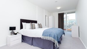 A modern bedroom with a bed, bedside table, and a view of a cityscape through large windows. - Clover Court by Aeria Apartments (London)