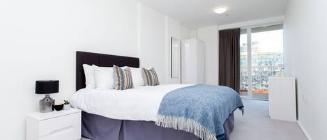 A modern bedroom with a bed, bedside table, and a view of a cityscape through large windows.