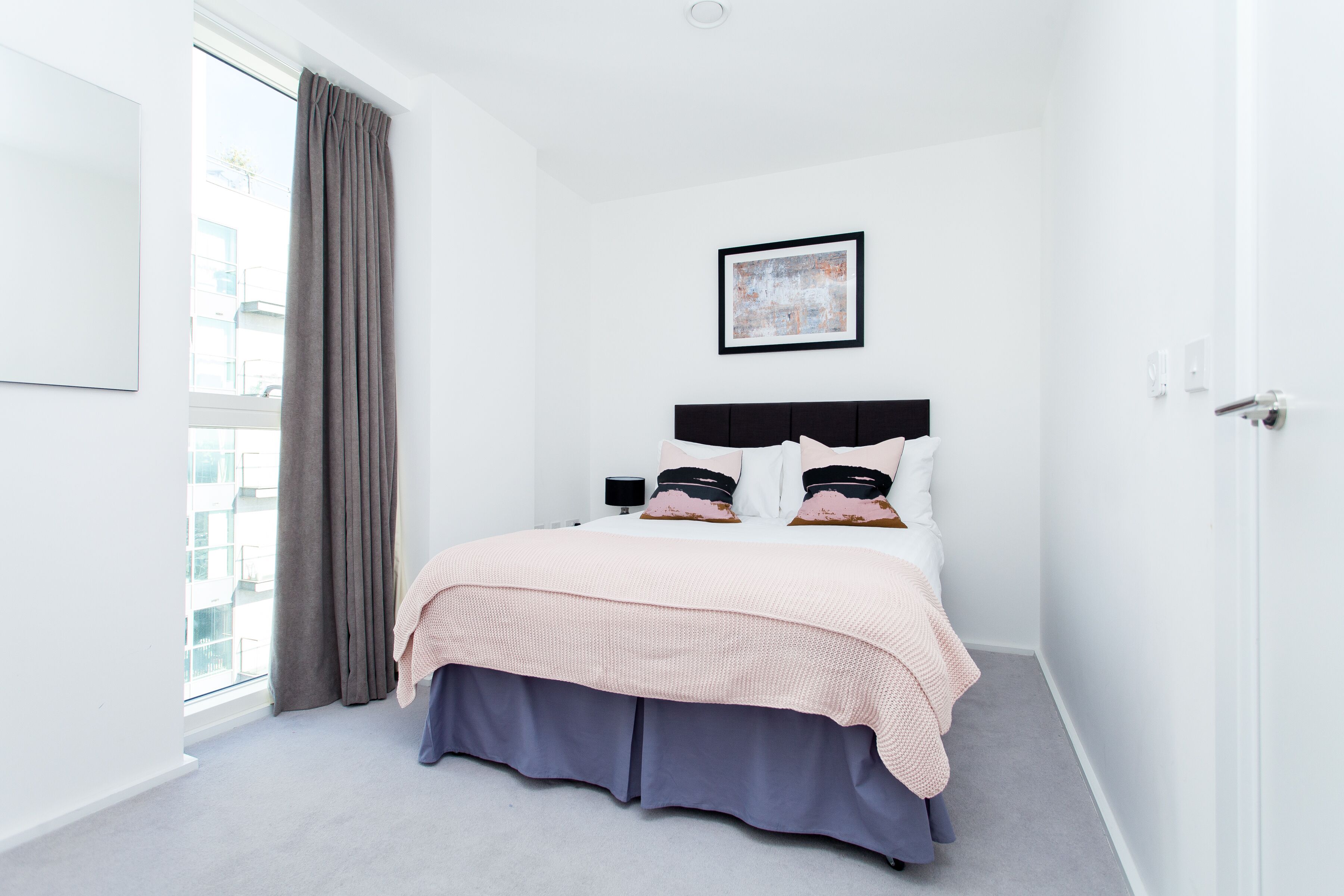 A hotel room with a bed, a window with curtains, a framed picture on the wall, and a door.