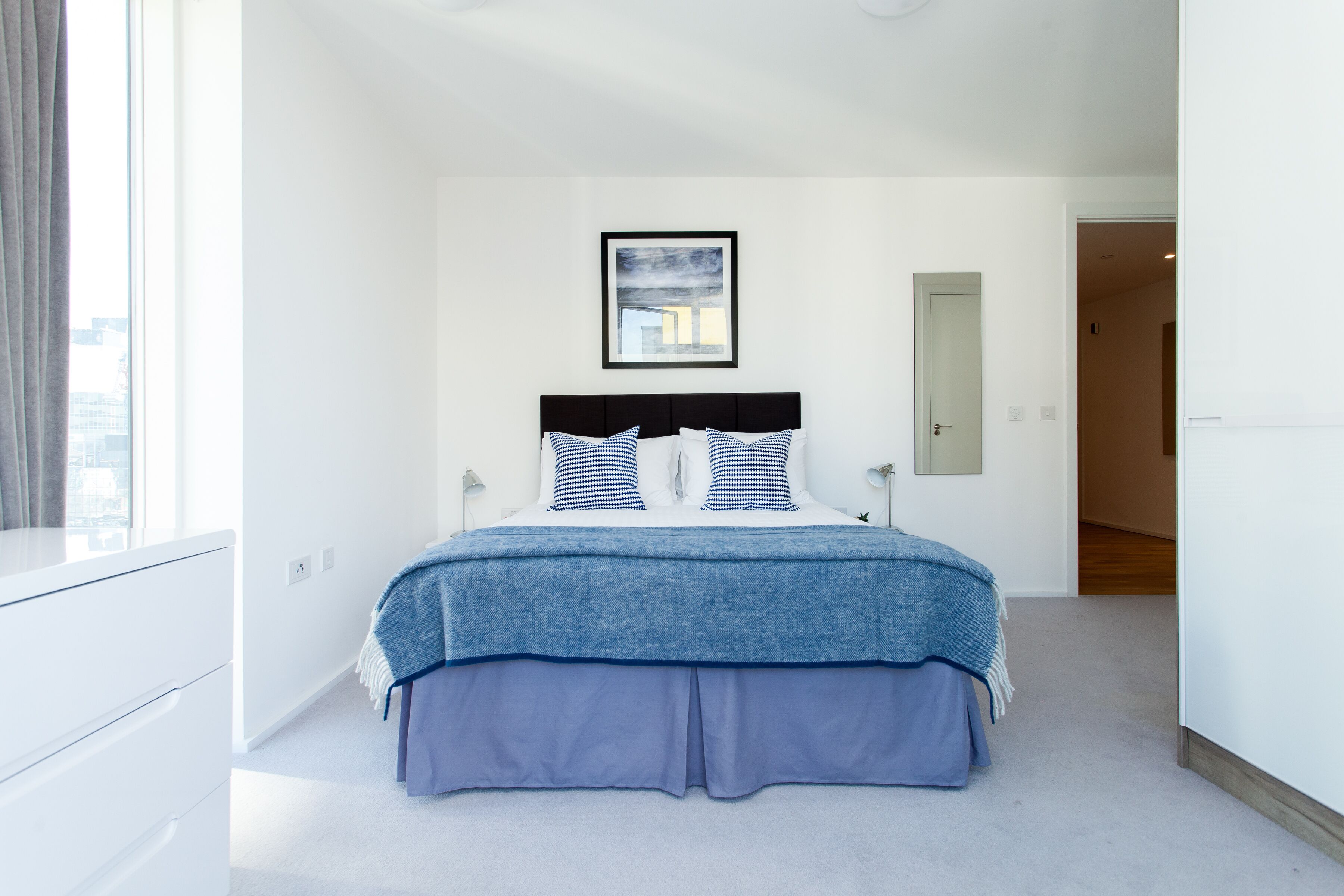 A bedroom with a bed, a window, a door, and a framed picture on the wall.