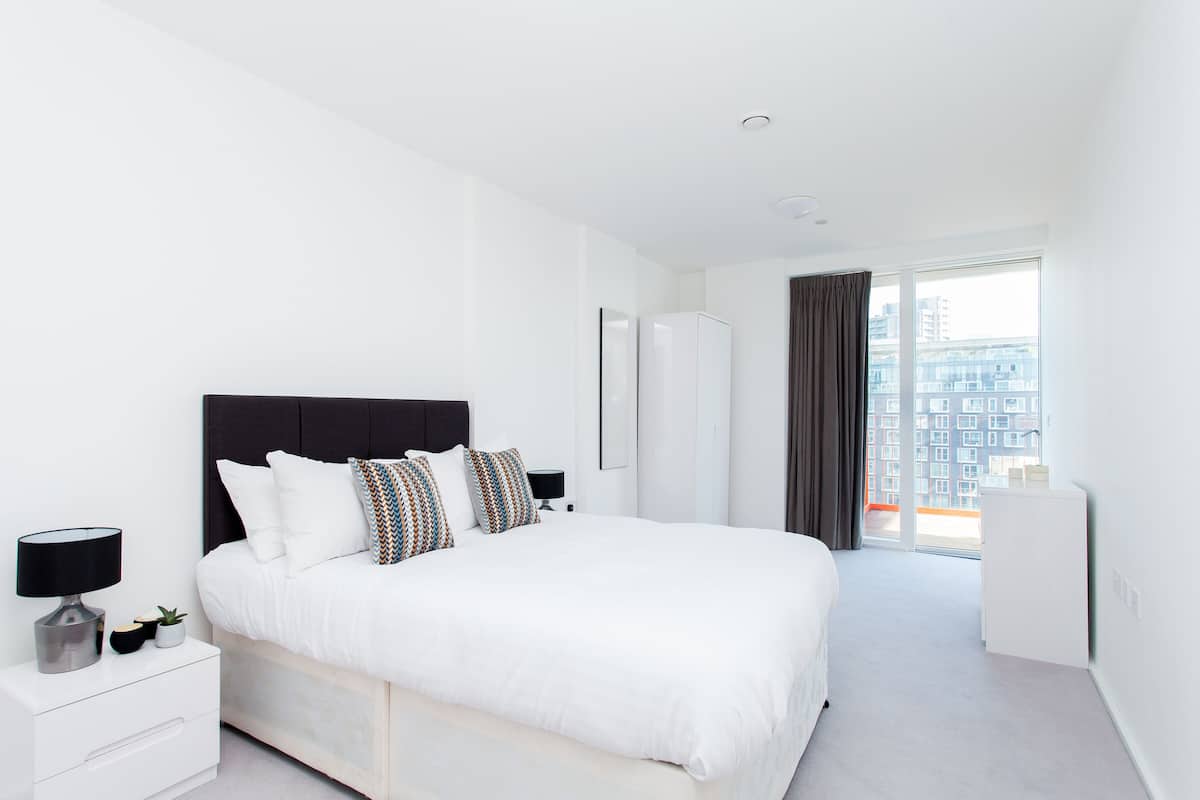Une chambre d’hôtel moderne avec un grand lit, des murs blancs, une tête de lit sombre, une table de chevet avec une lampe et une vue sur la ville par une grande fenêtre.