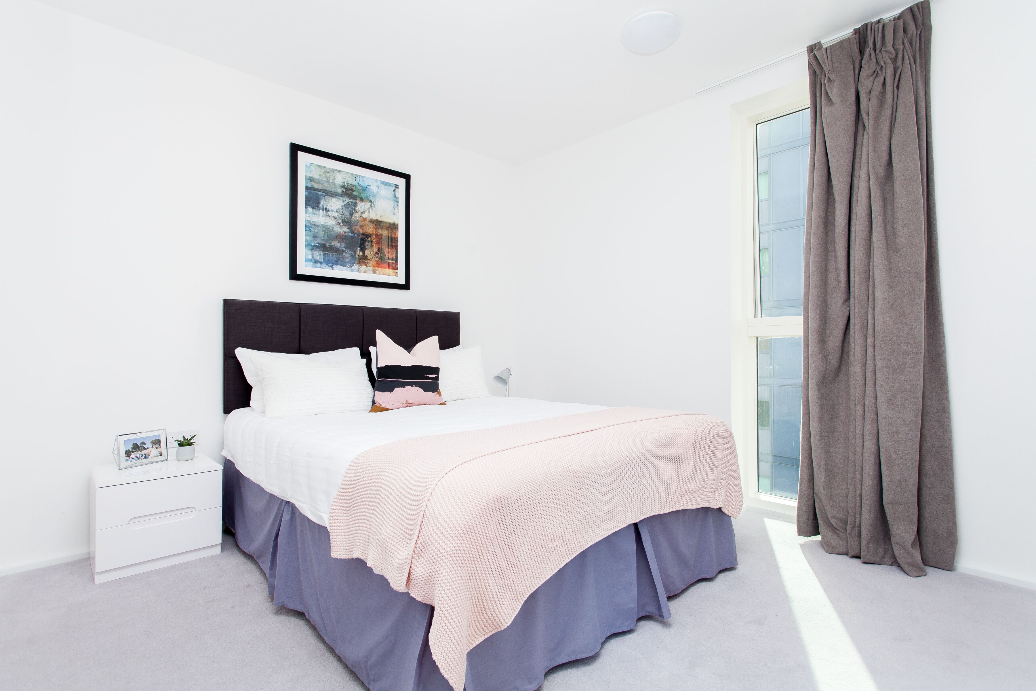 Een slaapkamer met een bed, nachtkastje, een ingelijste foto en een raam met gordijnen.
