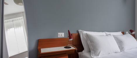 A neatly made bed with white linens, a wooden bedside table with a lamp, and a power outlet on the wall.