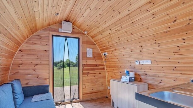 A cozy, semi-open wooden cabin with a sofa, a small kitchenette, and a view of a grassy field. - Cosy Camping Suffolk (Ipswich)