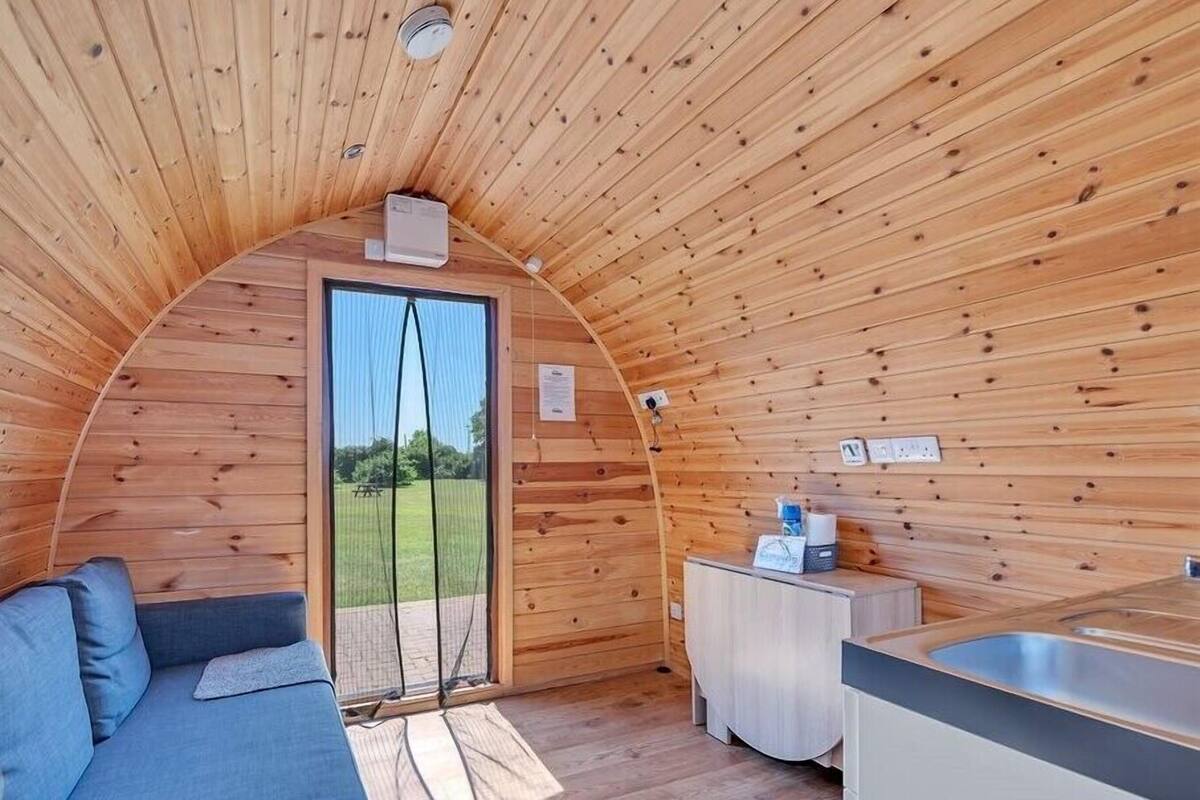 Une cabane en bois accueillante, semi-ouverte, avec un canapé, une petite kitchenette et une vue sur un champ verdoyant.