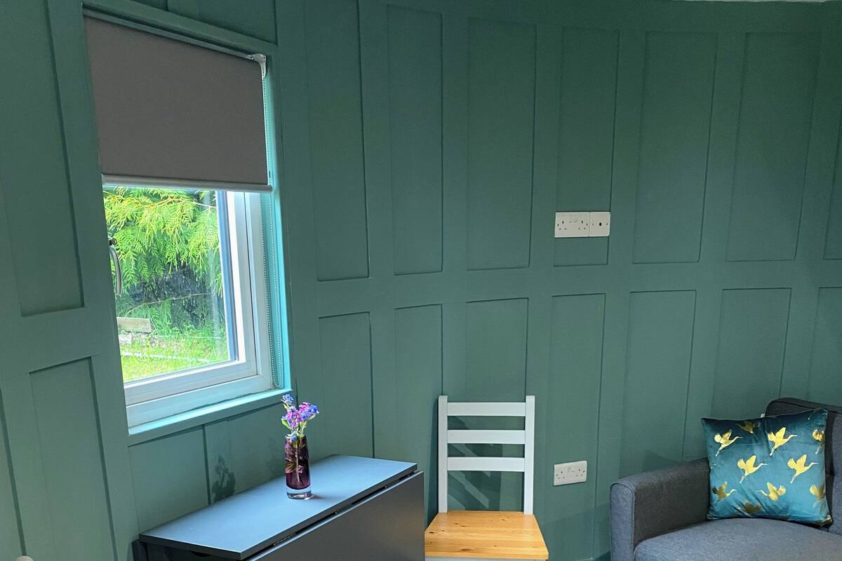 Une pièce aux murs de couleur turquoise, avec un bureau et une chaise, un canapé et une fenêtre donnant sur un paysage verdoyant.