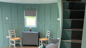 A room with a wooden floor, a grey sofa, two wooden chairs, a small table, a window with a view, and a spiral staircase. - Cosy Camping Suffolk (Ipswich)