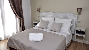 Una cama bien hecha con ropa blanca y una toalla doblada, dos mesitas de noche con lámparas y una ventana con cortinas.