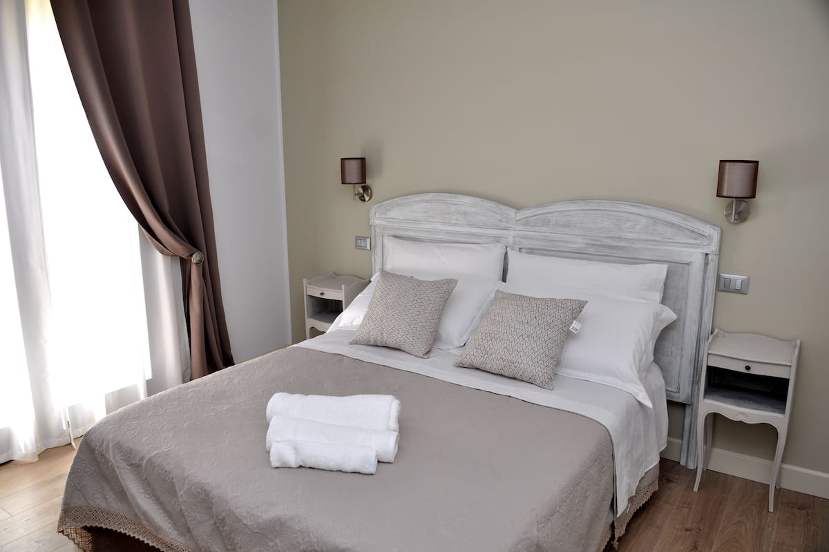 Un lit bien fait, avec du linge de lit blanc et une serviette pliée, deux tables de chevet avec des lampes et une fenêtre avec des rideaux.